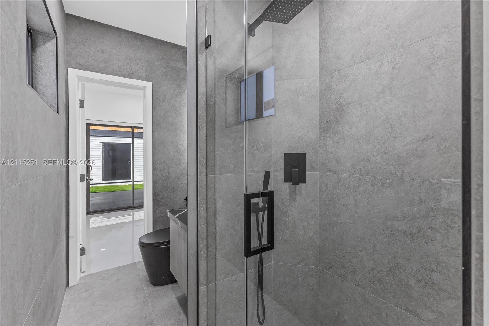 Walk-in glass-enclosed shower featuring large-format gray tile, a rainfall showerhead, and matte black fixtures, complemented by a floor-mounted toilet and a gray vanity cabinet