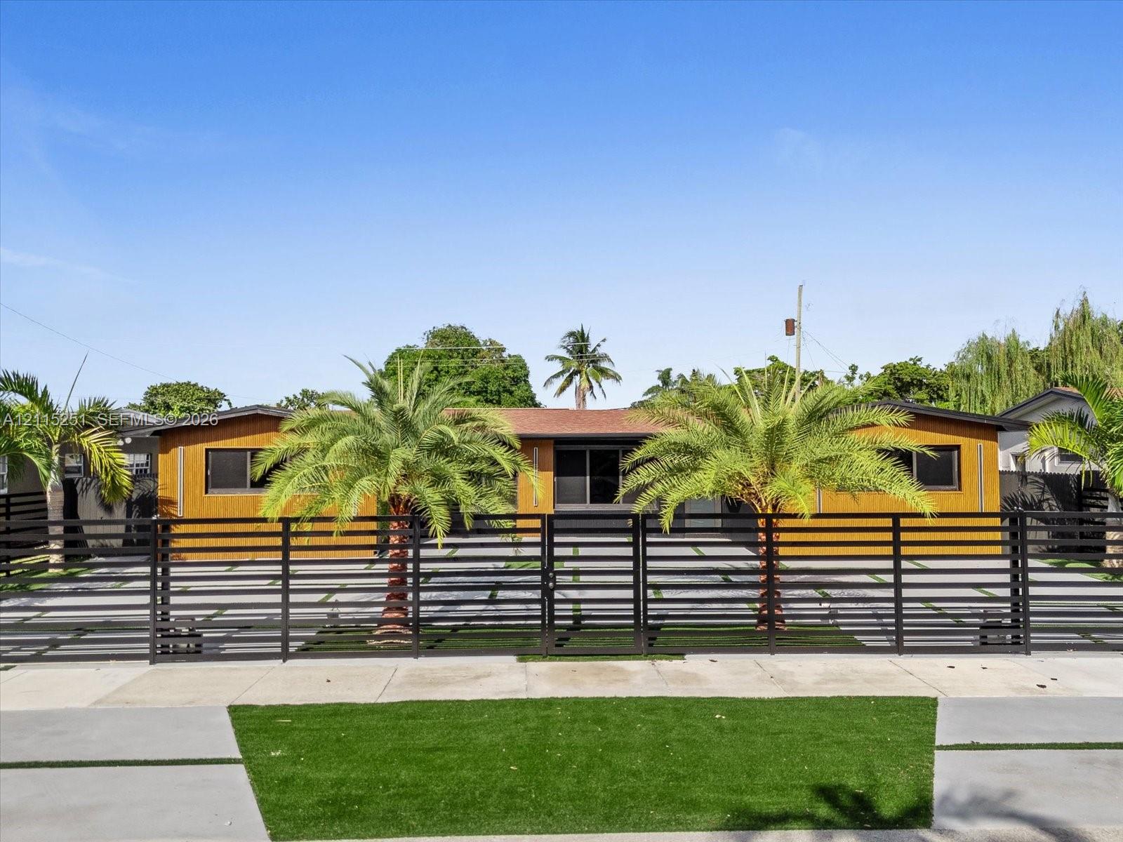 Single-story exterior featuring wood-finish paneling, horizontal slat metal fencing, paved driveway with grass inserts, and integrated palm trees