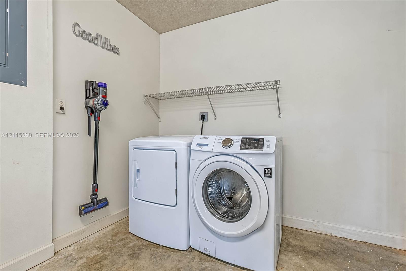 Laundry area featuring white washer and dryer units, a wall-mounted wire shelf, and exposed concrete flooring