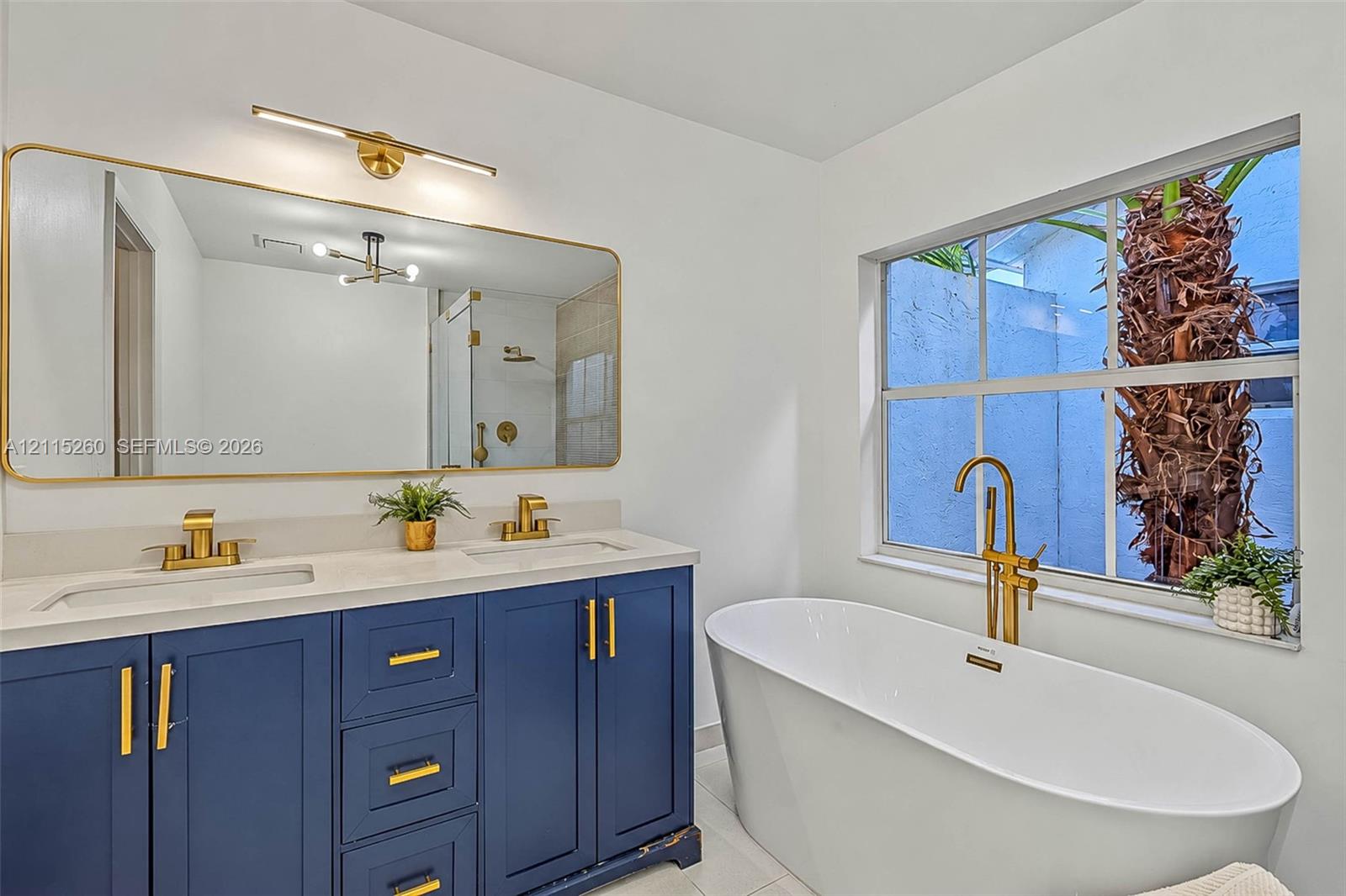 Dual-sink vanity with navy blue cabinetry, gold-tone hardware, and a quartz countertop, paired with a freestanding soaking tub featuring a gold-tone floor-mounted faucet and an expansive gold-rimmed mirror