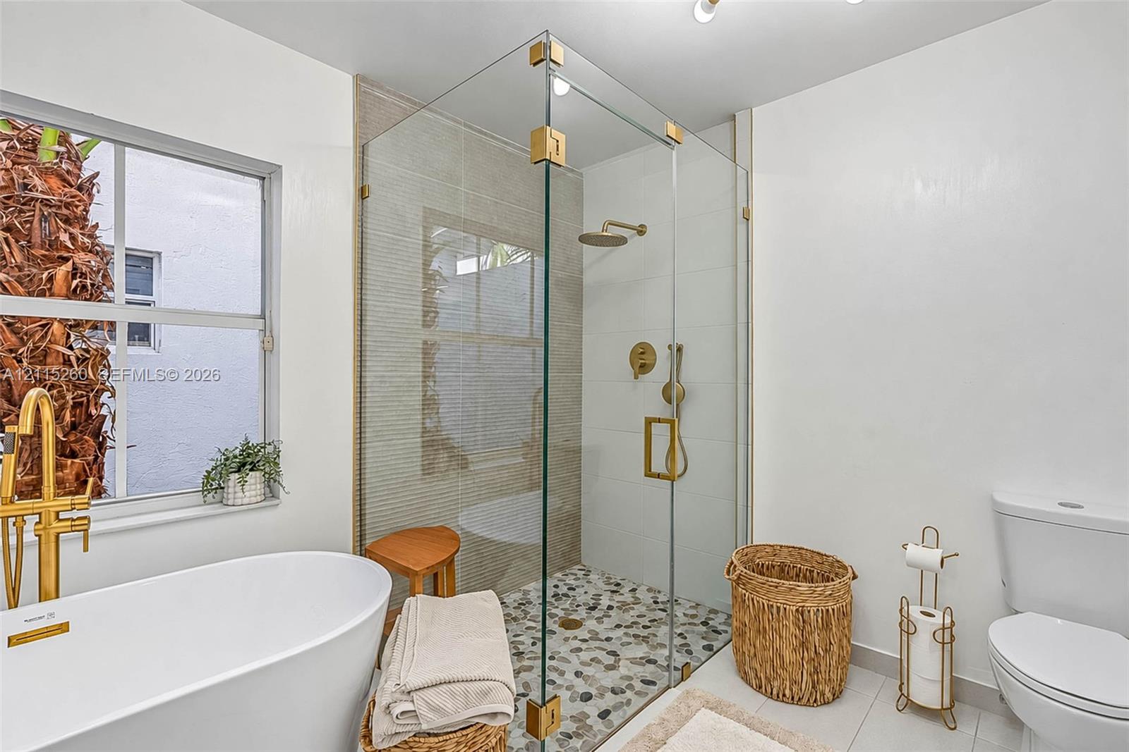 Freestanding soaking tub with a brass-finish floor-mounted faucet, frameless glass-enclosed shower with a pebble stone floor, and brass-finish shower fixtures