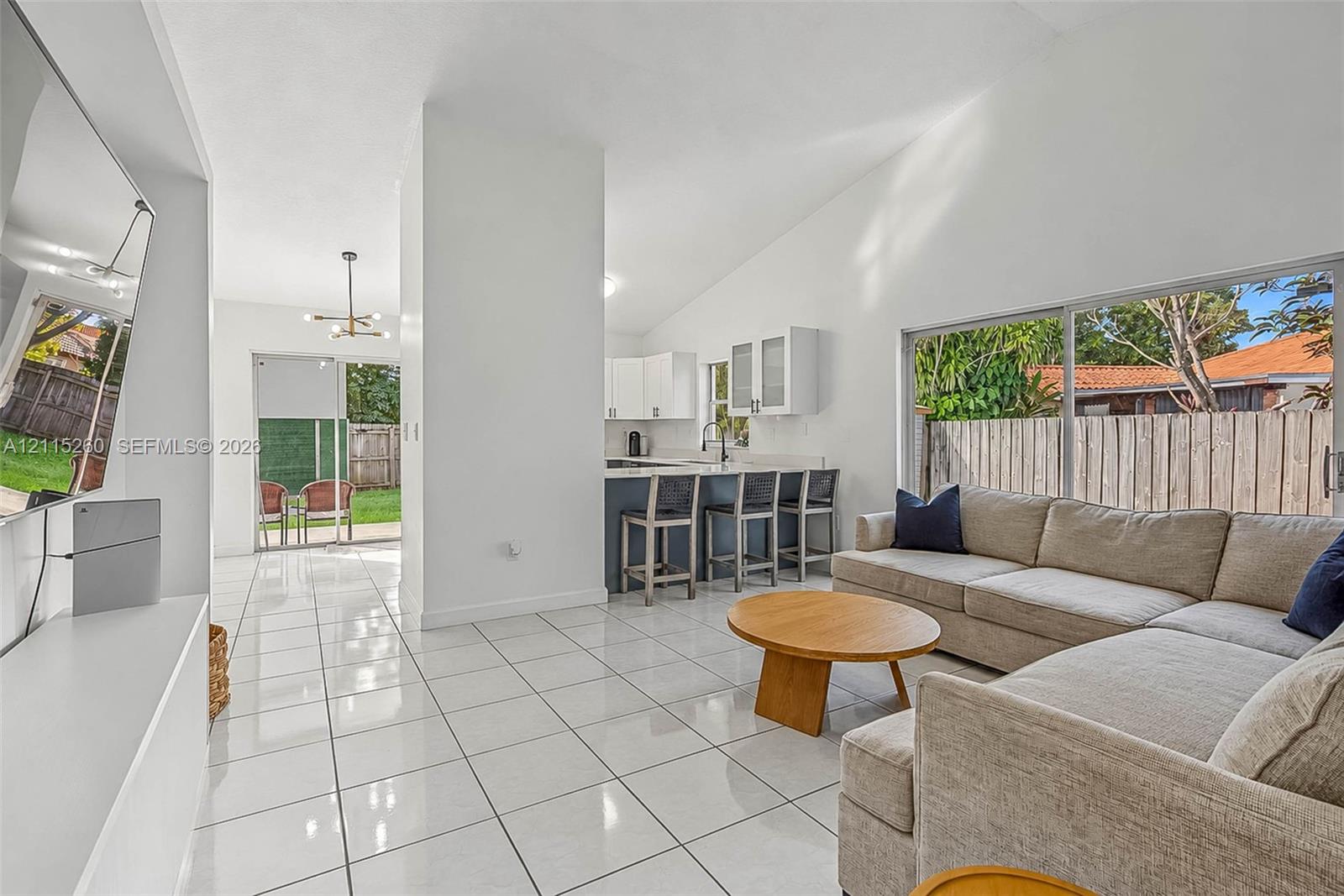 Open-concept living space featuring vaulted ceilings, large-format tile flooring, sliding glass patio doors, and a kitchen equipped with a breakfast bar and white upper cabinetry