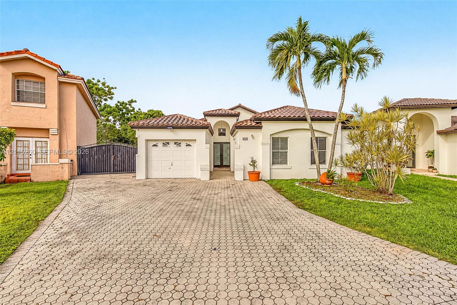 Stucco exterior with a tiled roof, paver driveway, attached garage, and landscaping featuring mature palms
