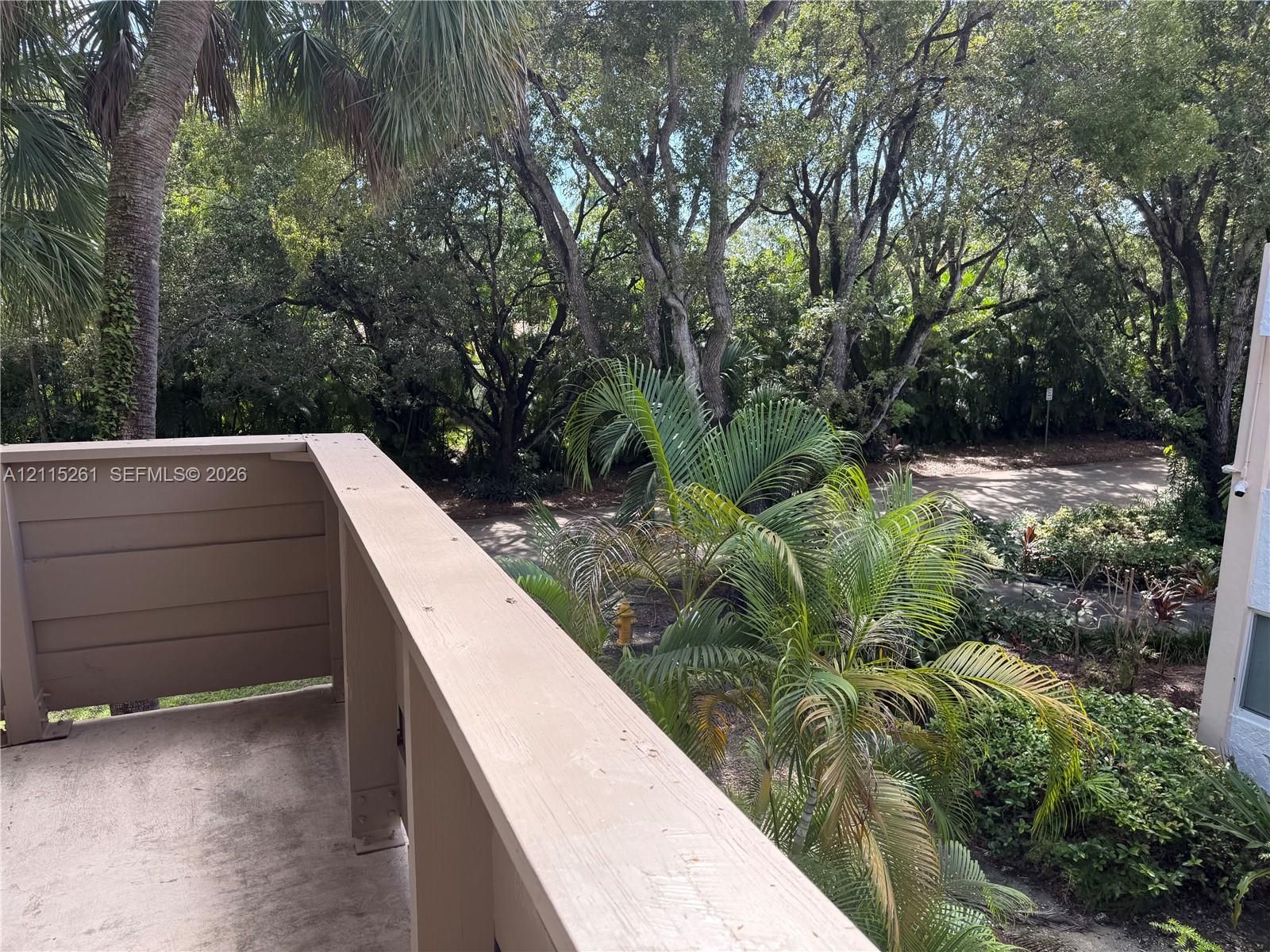 Covered balcony featuring wood-tone railings and concrete flooring, overlooking mature trees and lush landscaping