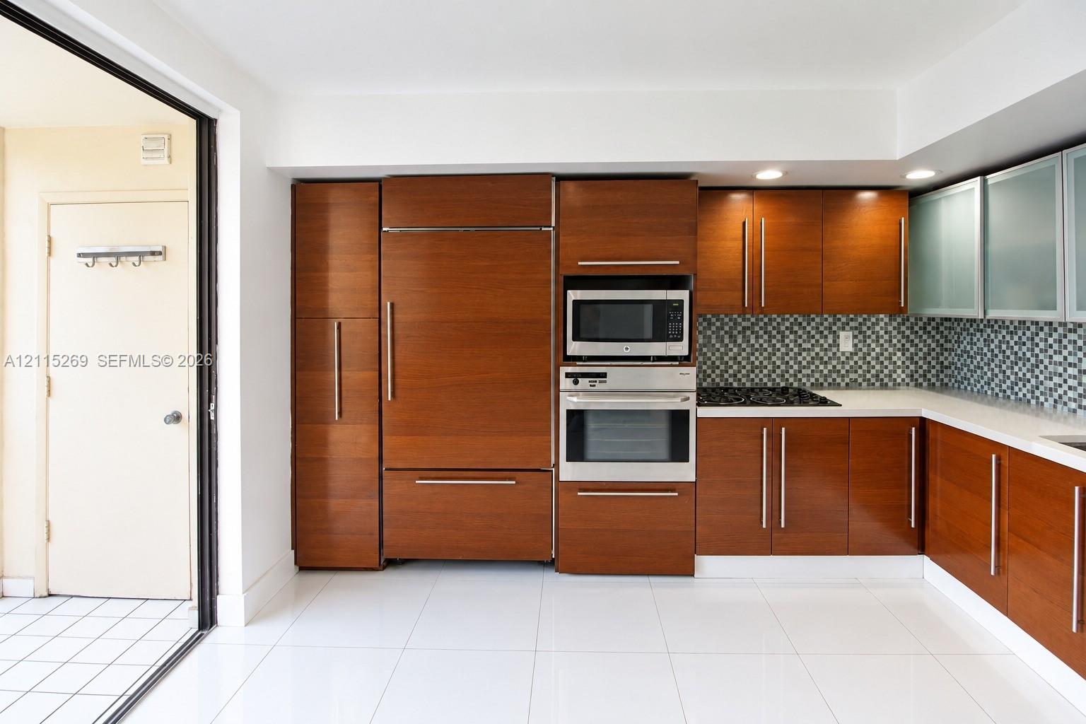 Kitchen featuring custom wood-finish cabinetry with integrated appliances, stainless steel wall-mounted oven and microwave, mosaic tile backsplash, solid-surface countertops, and large-format tile flooring