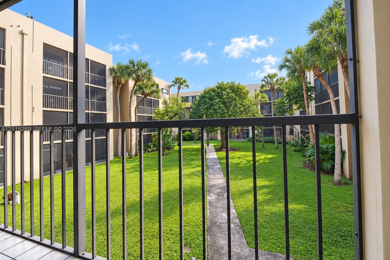 Screened balcony with metal railing overlooking a landscaped courtyard featuring a lawn, mature palm trees, and a paved walkway