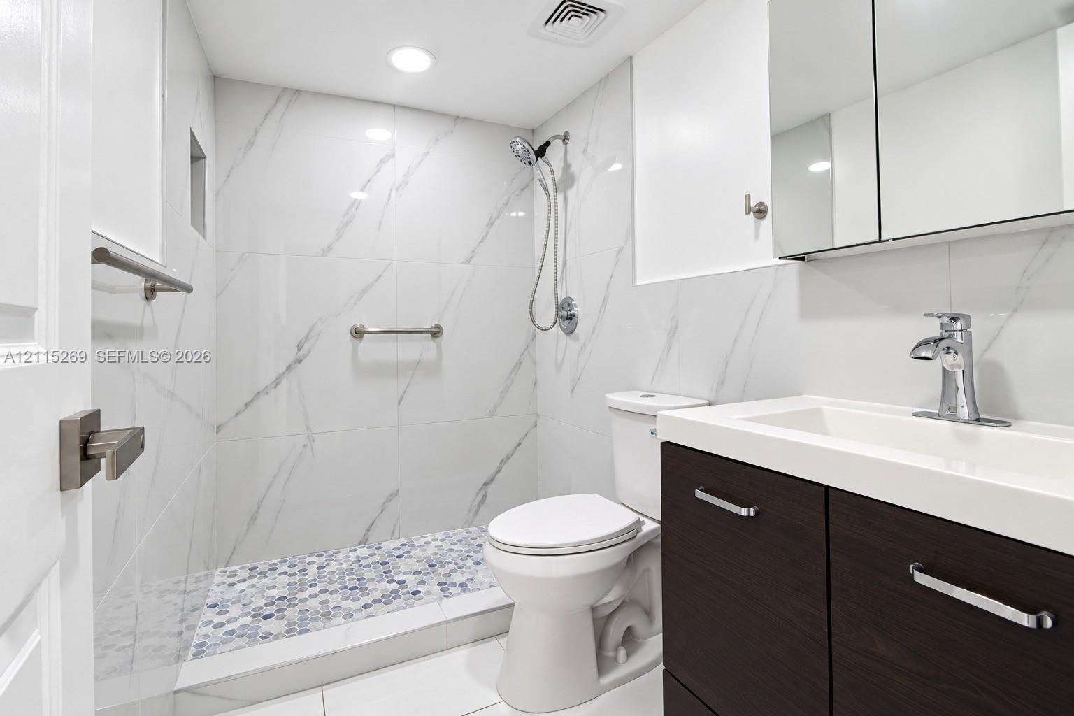 Bathroom featuring white wall tile with stone-pattern veining, a walk-in shower with penny-round floor tile, a dark wood-finish vanity with an integrated basin, and a mirrored medicine cabinet