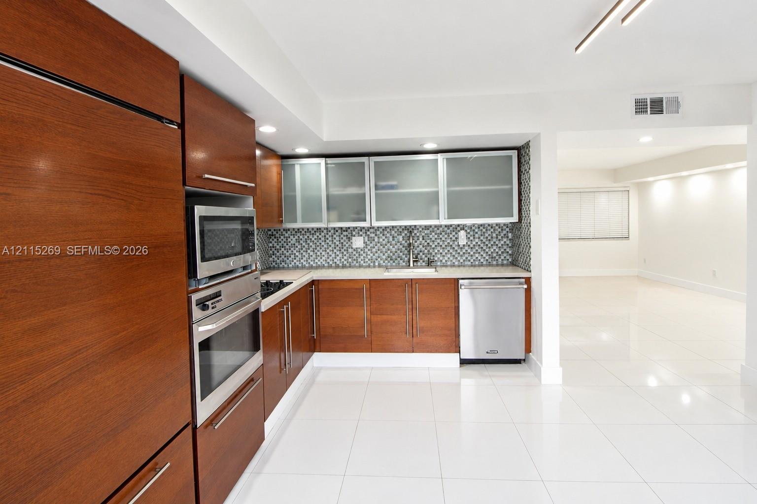 Kitchen featuring wood-finish cabinetry, stainless steel built-in oven and microwave, patterned tile backsplash, frosted-glass upper cabinet inserts, and polished tile flooring
