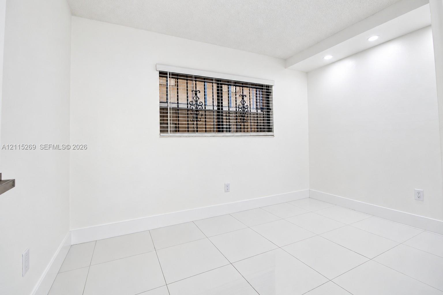Bright room featuring white tile flooring, recessed ceiling lighting, and a window equipped with horizontal blinds and decorative metal security grilles
