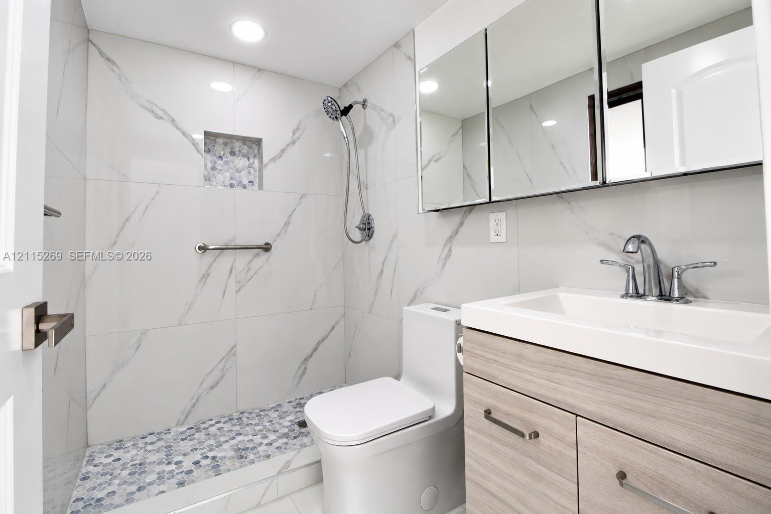 Walk-in shower with stone-finish tile surround, integrated shower niche, penny-tile floor, wood-finish vanity, and recessed medicine cabinet