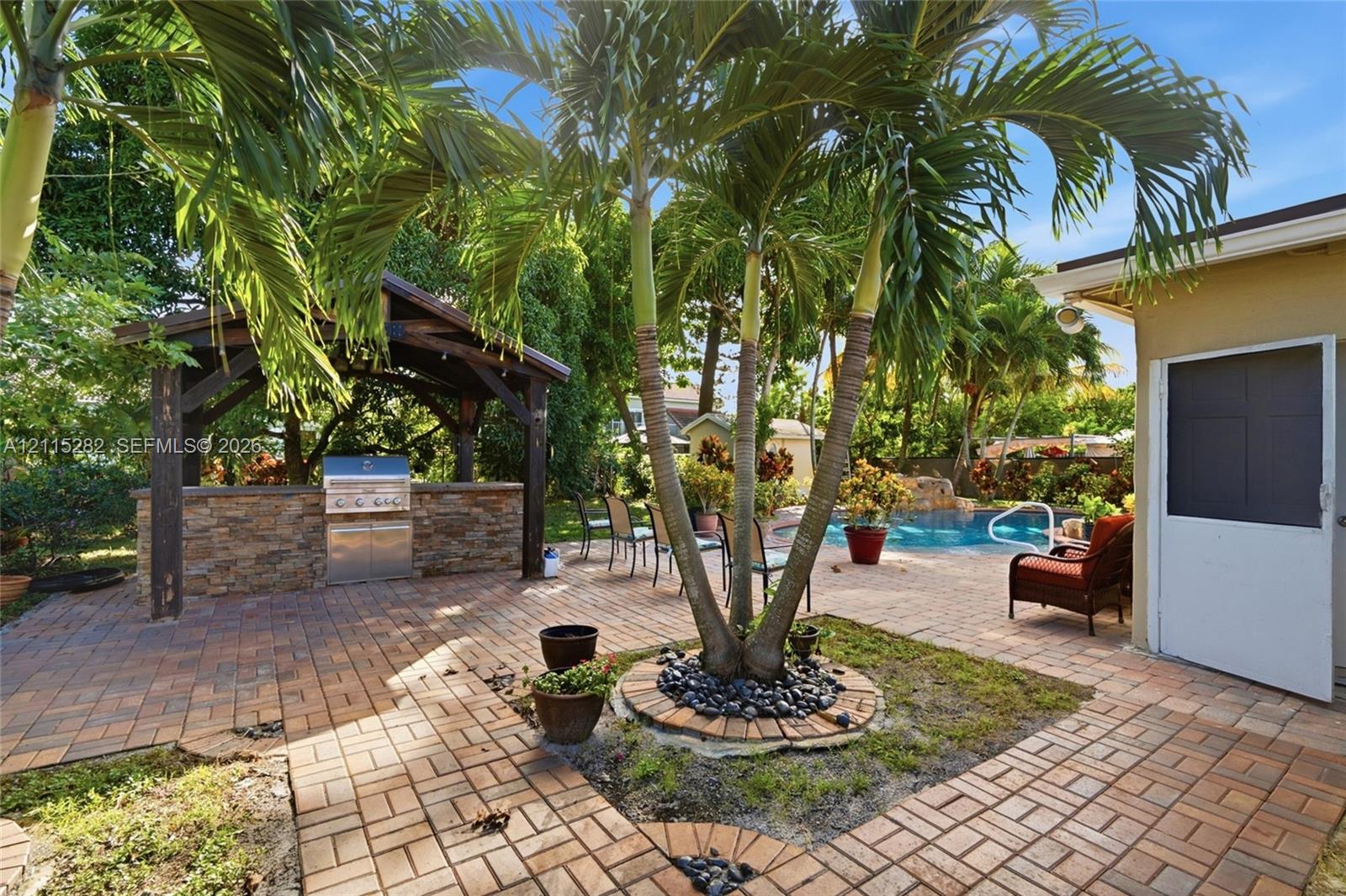 Outdoor living space featuring a brick-paved patio, a covered pavilion with a built-in stone-finish grill station, a swimming pool, and mature palm trees