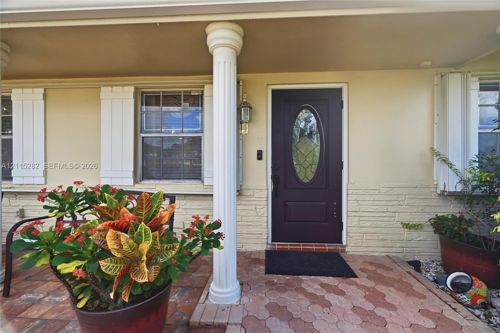 Covered entryway featuring a fluted column, stone-finish exterior wall, paver flooring, and a central entry door with a decorative glass insert Covered entryway featuring a fluted column, stone-finish exterior wall, paver flooring, and a central entry door with a decorative glass insert