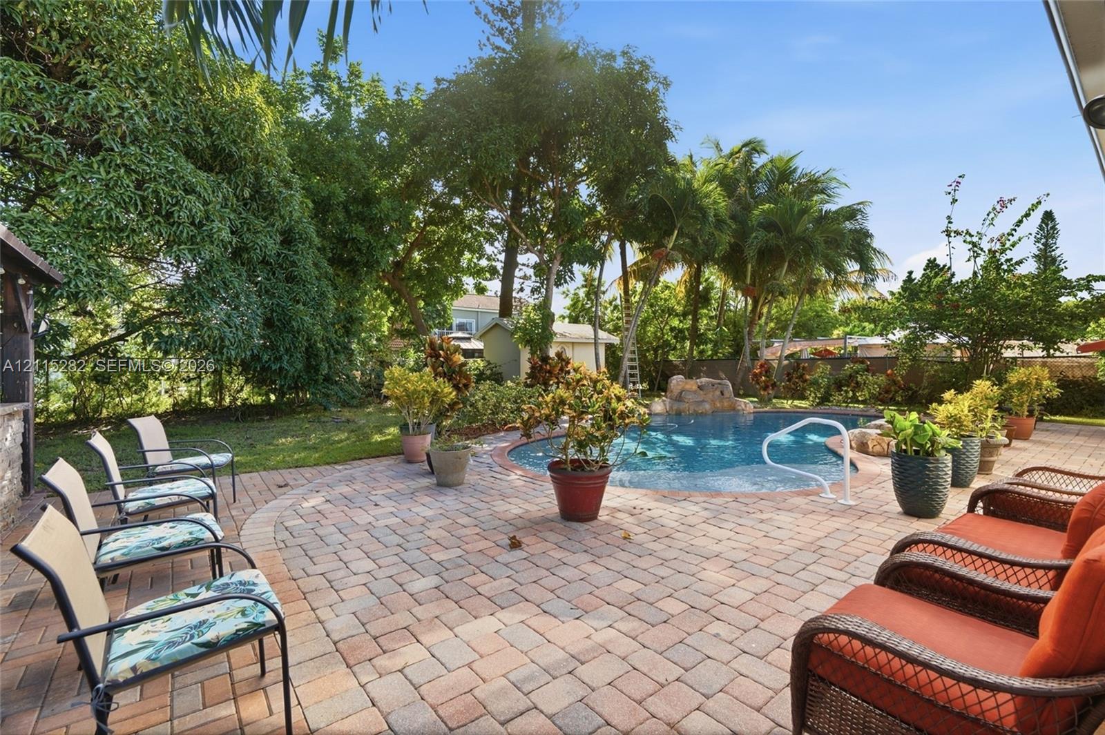 Paver patio area featuring an in-ground swimming pool with a rock water feature, surrounded by mature trees and lush landscaping