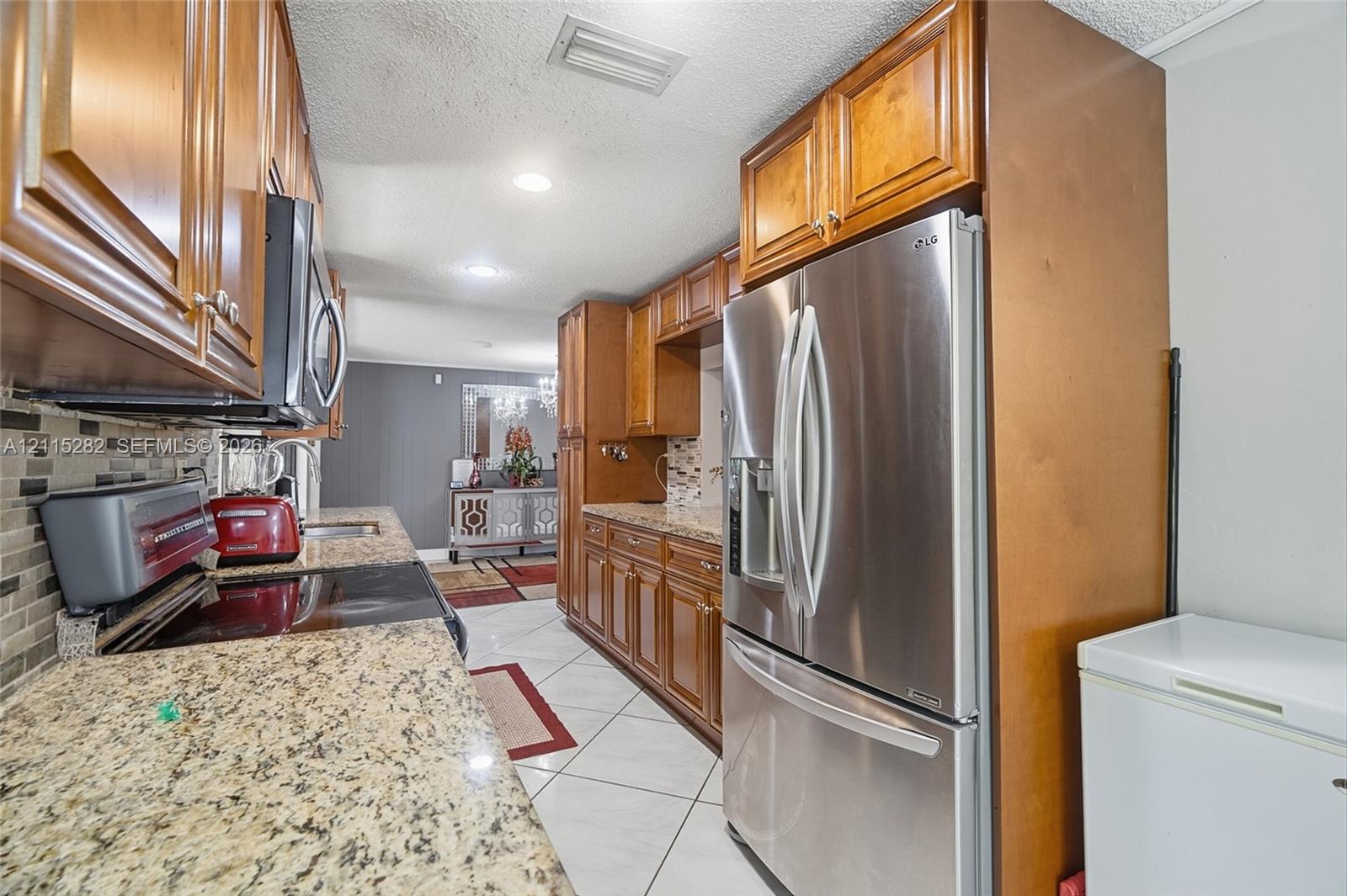 Kitchen featuring wood cabinetry, stone countertops, stainless steel refrigerator, mosaic tile backsplash, and tile flooring