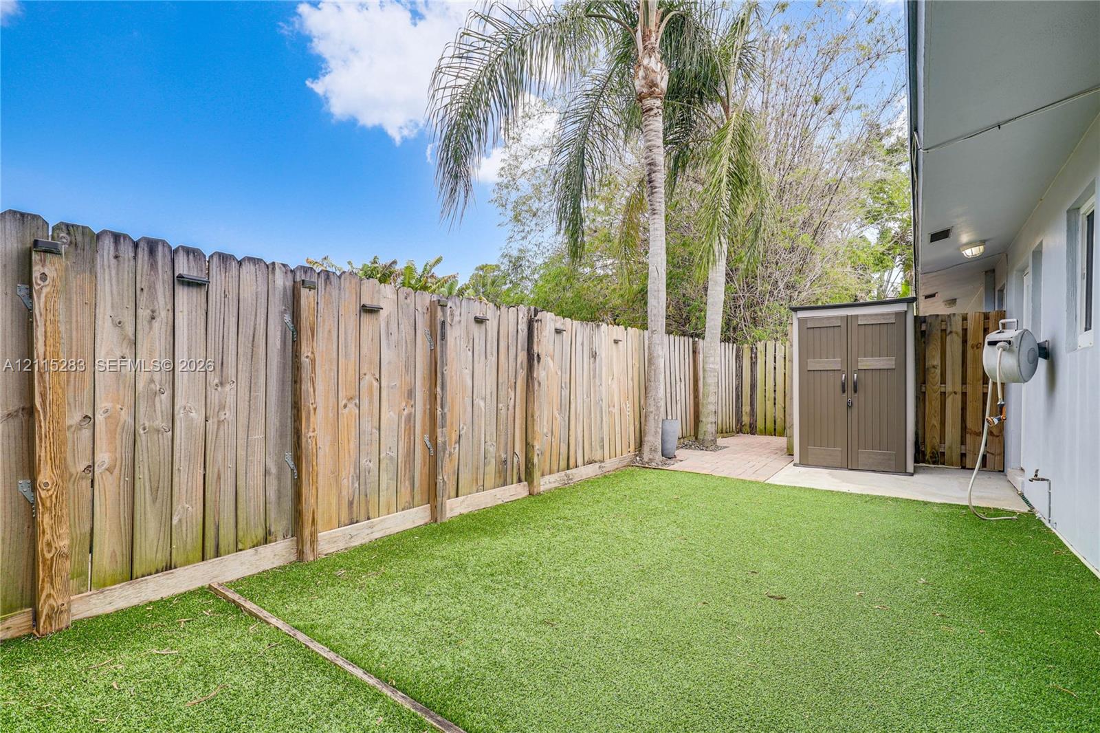 Private side yard featuring a wood privacy fence, artificial turf, brick-paver patio area, and mature palm trees
