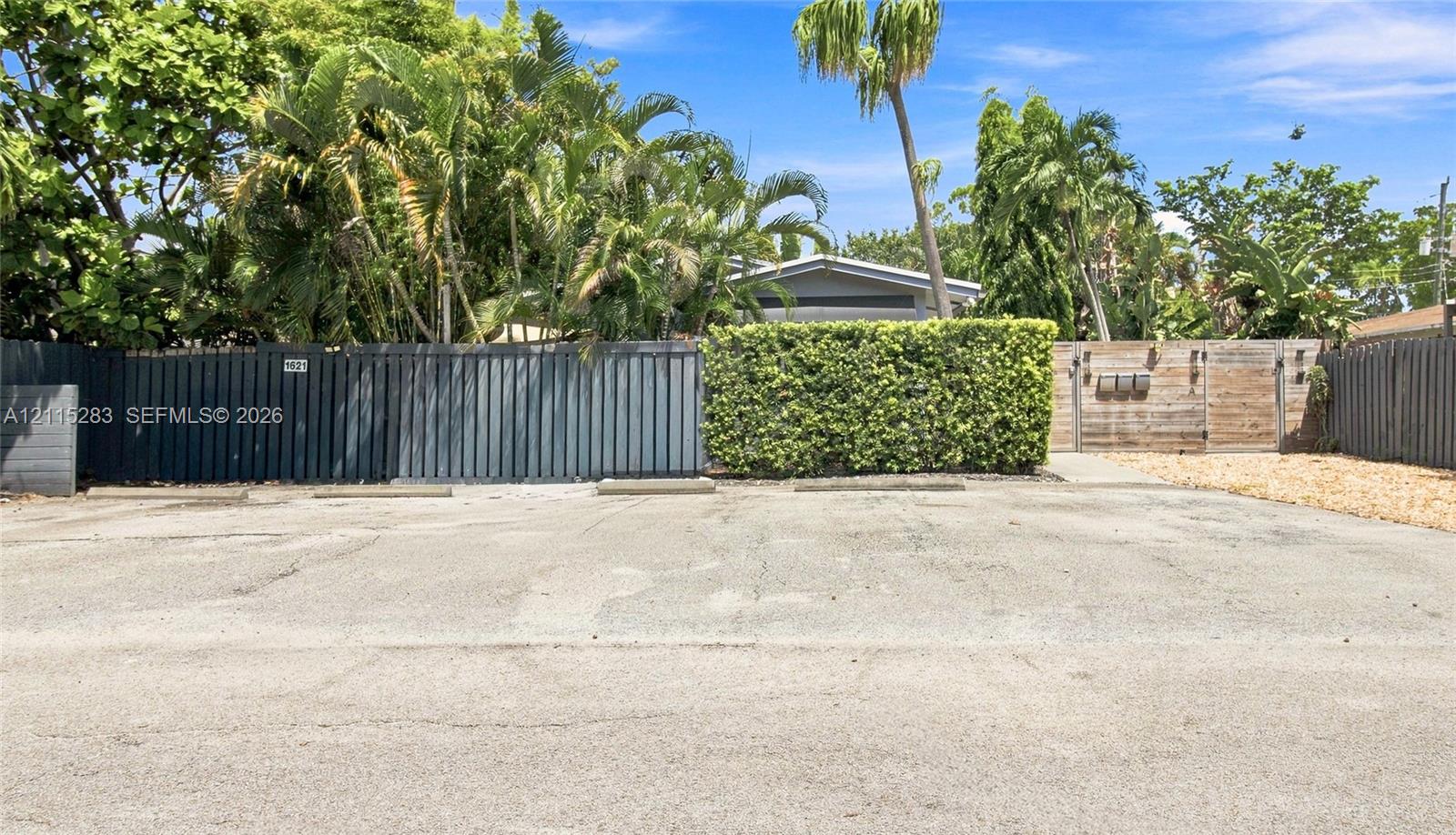 Paved off-street parking area featuring concrete wheel stops, dark-stained horizontal wood privacy fencing, and mature tropical landscaping
