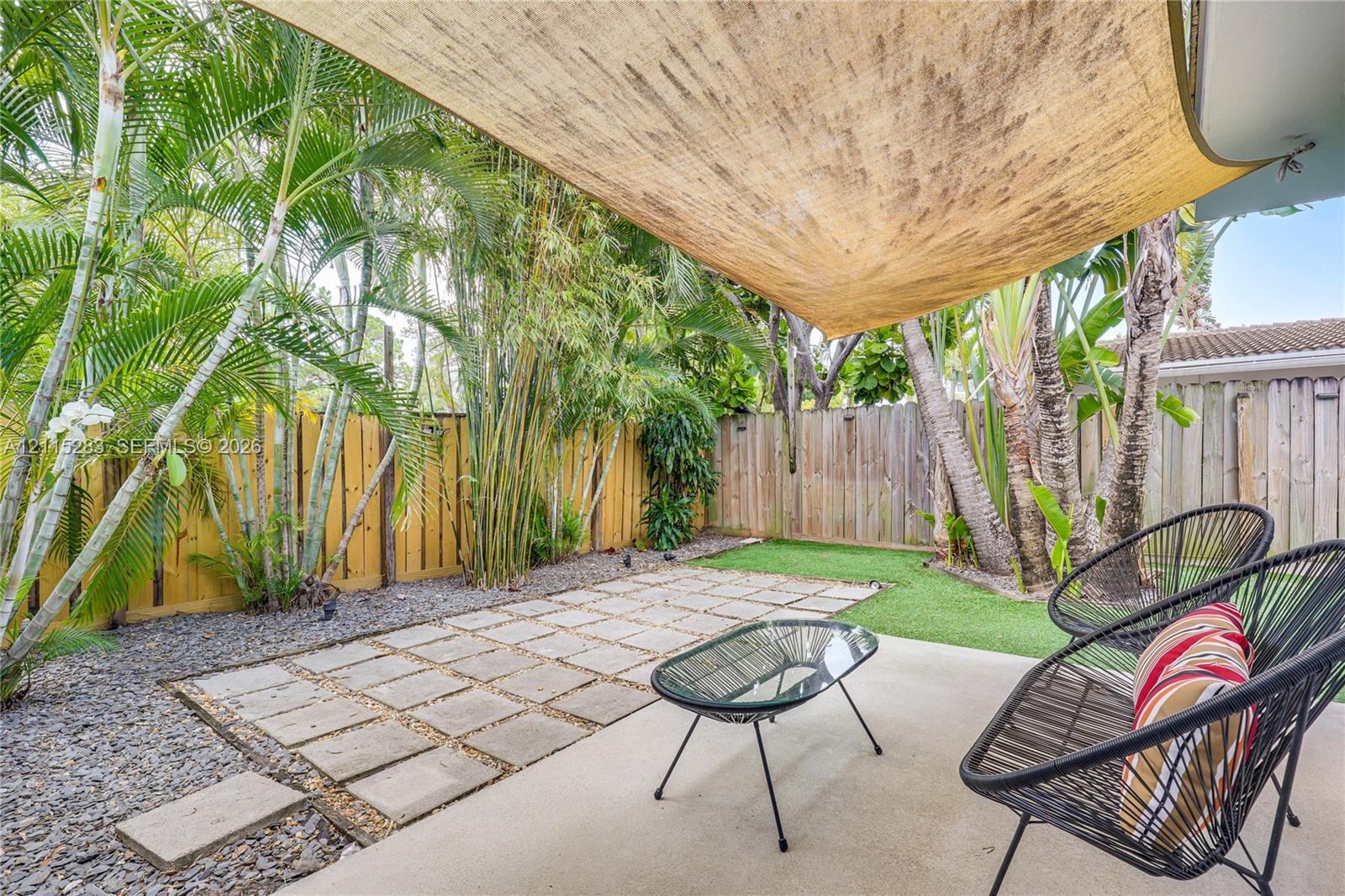 Covered concrete patio featuring a fabric shade sail, paver stone hardscaping, artificial turf, perimeter wood fencing, and mature palm landscaping