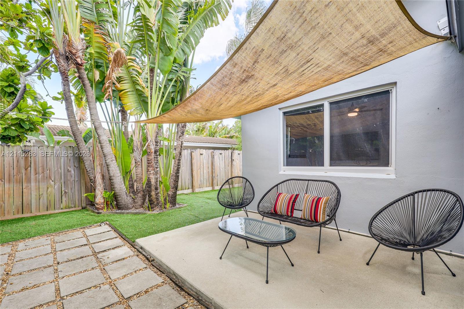 Backyard featuring a concrete patio, synthetic turf lawn, wood privacy fencing, mature palm trees, and a tension-mounted shade sail