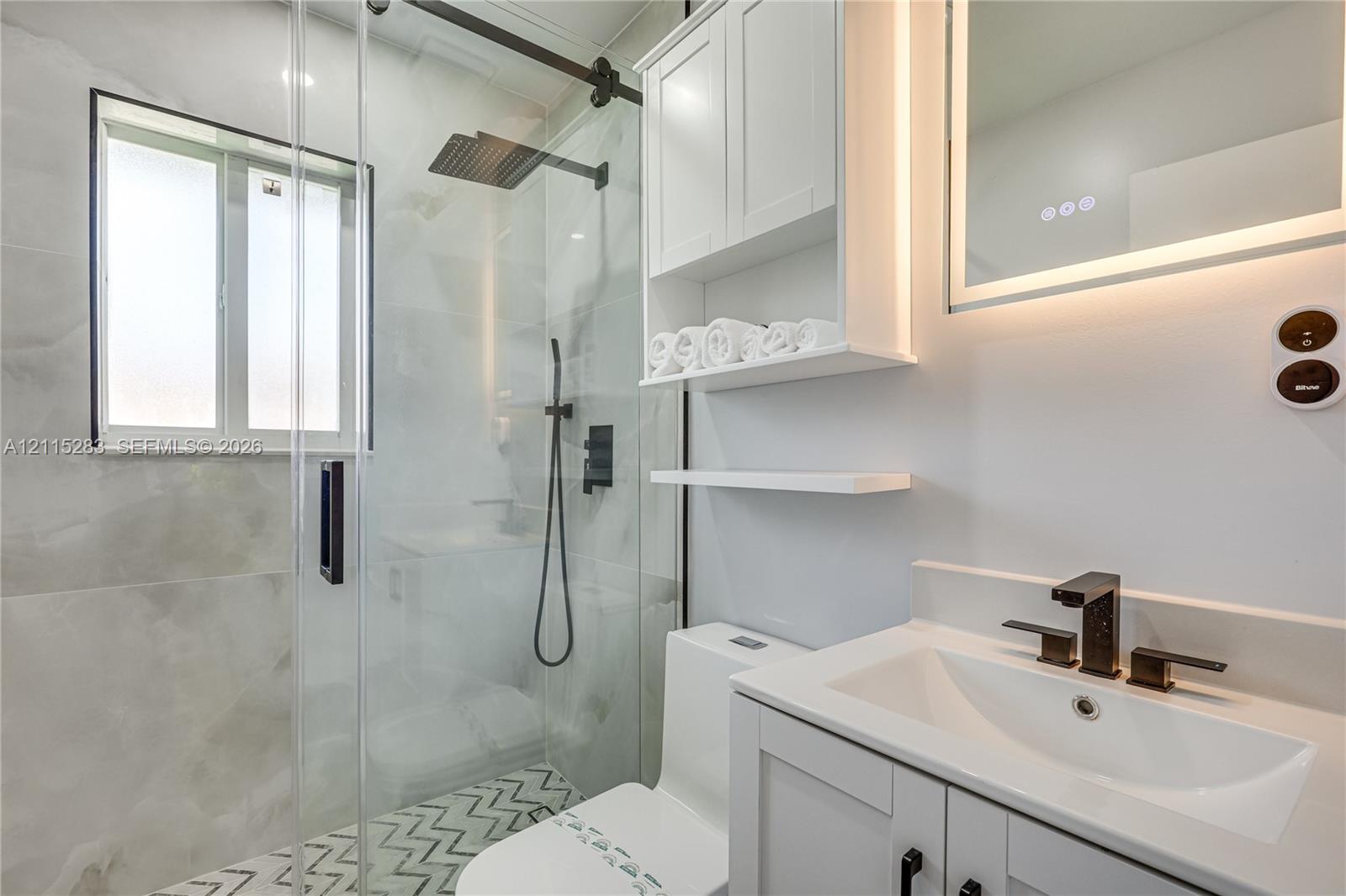 Bathroom featuring a frameless glass shower enclosure with matte black fixtures, rainfall showerhead, custom built-in white cabinetry, and geometric patterned floor tiling