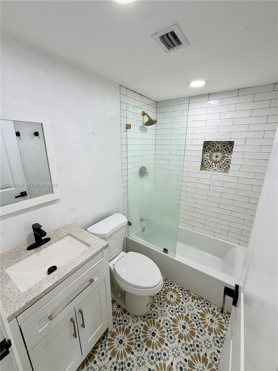 Bathroom featuring patterned tile flooring, white subway tile shower surround with a decorative niche, frameless glass shower panel, and a stone-topped vanity with matte black fixtures