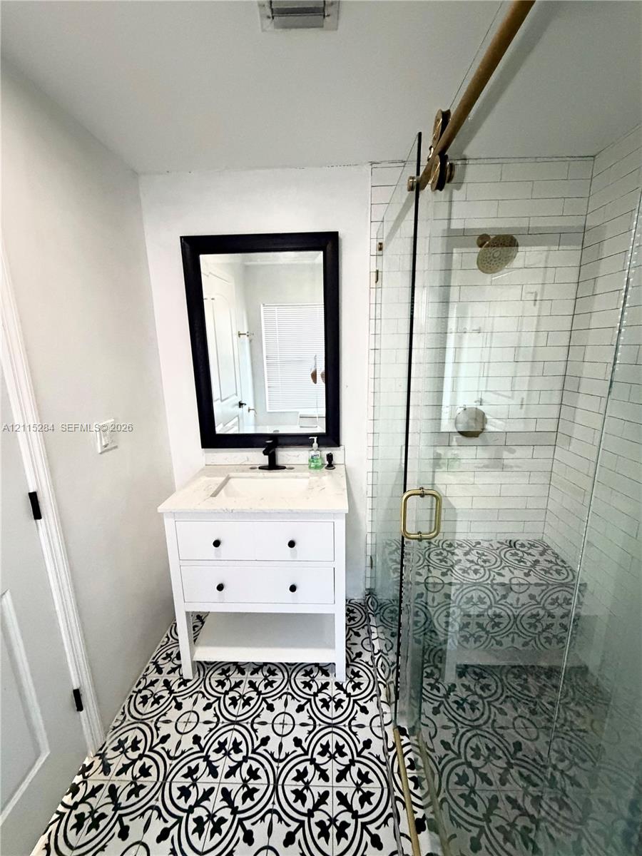 White vanity with a stone countertop, framed mirror, glass-enclosed shower with white subway tile walls, and decorative patterned floor tiling