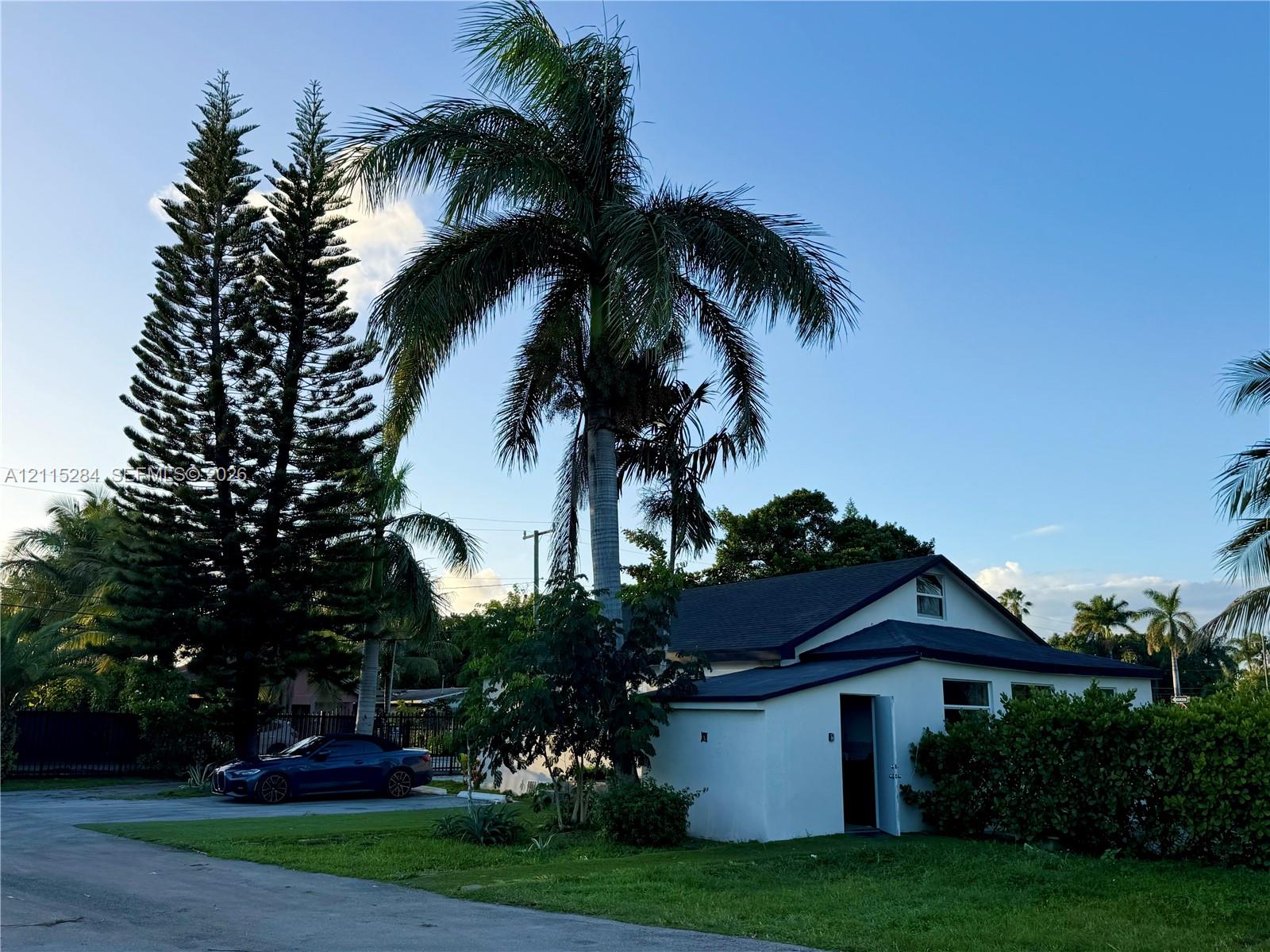 Single-story exterior featuring a gabled roofline, light-colored smooth finish siding, and mature tropical landscaping including tall pine and palm trees