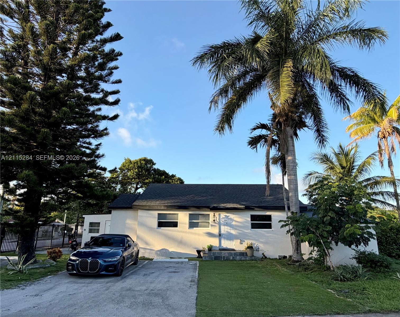 Single-story exterior featuring a light-colored stucco facade, shingled roof, concrete driveway, and mature palm and pine landscaping