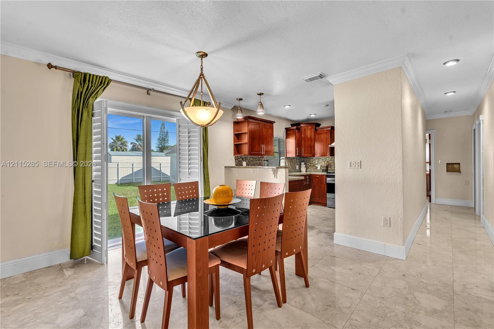 Dining area featuring stone-finish tile flooring, recessed lighting, and a geometric pendant chandelier, accompanied by a kitchen with wood-finish cabinetry and sliding glass doors