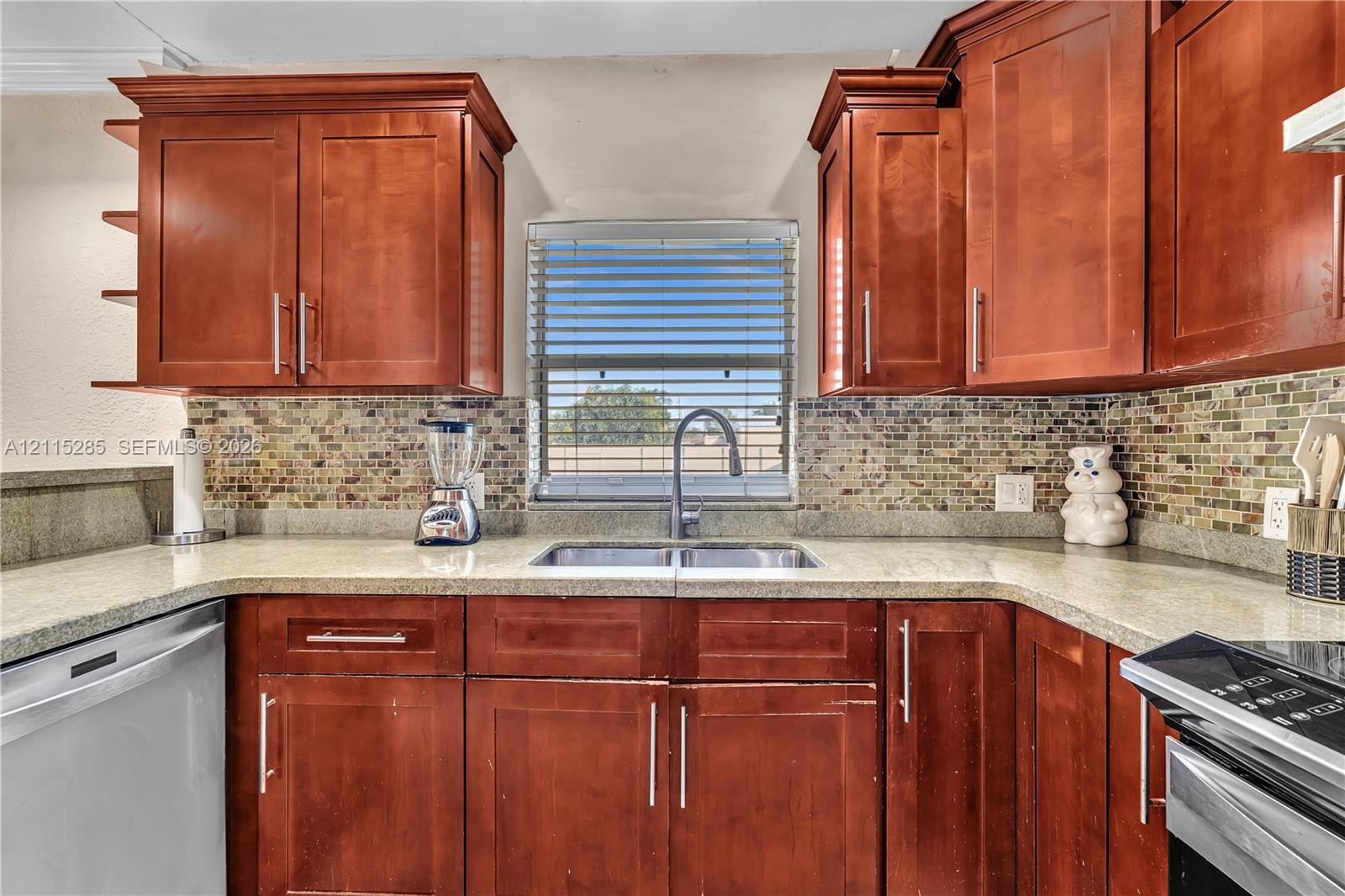 Kitchen featuring wood cabinetry, stone-surface countertops, mosaic tile backsplash, dual-basin under-mount sink, and stainless steel hardware
