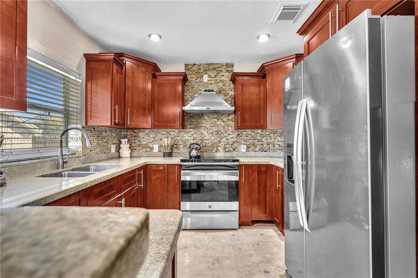 Kitchen featuring warm-toned wood cabinetry, mosaic tile backsplash, stainless steel appliances, stone-finish countertops, and recessed lighting