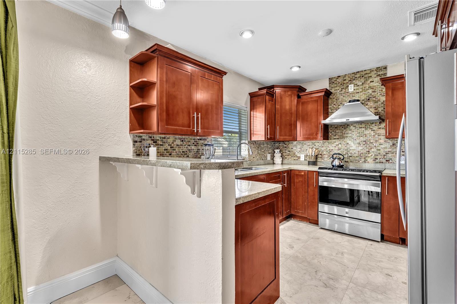 Kitchen featuring warm-tone cabinetry, stone-finish countertops, stainless steel appliances, a mosaic tile backsplash, and tile flooring