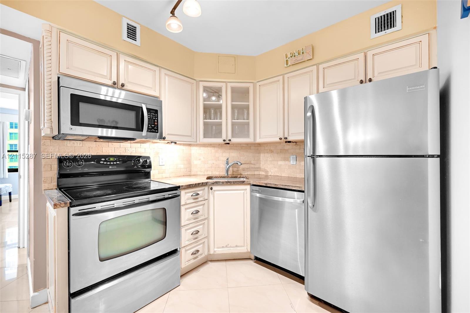 Kitchen featuring stainless steel appliances, light-toned cabinetry with glass-front display panels, tiled backsplash, and stone-finish countertops