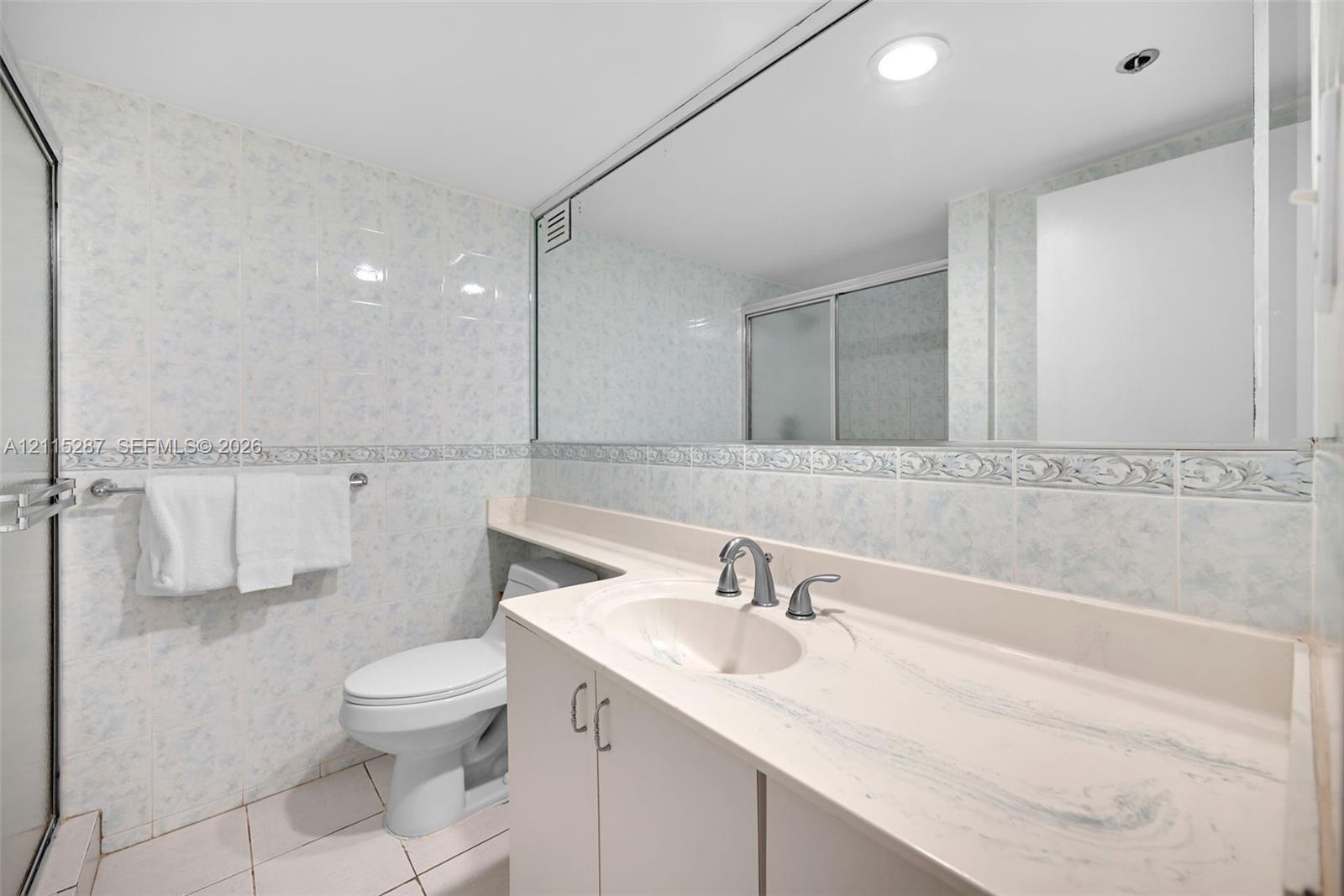 Bathroom featuring wall-to-wall mirror, solid-surface vanity countertop with integrated sink, floor-to-ceiling ceramic tile, and glass-enclosed shower