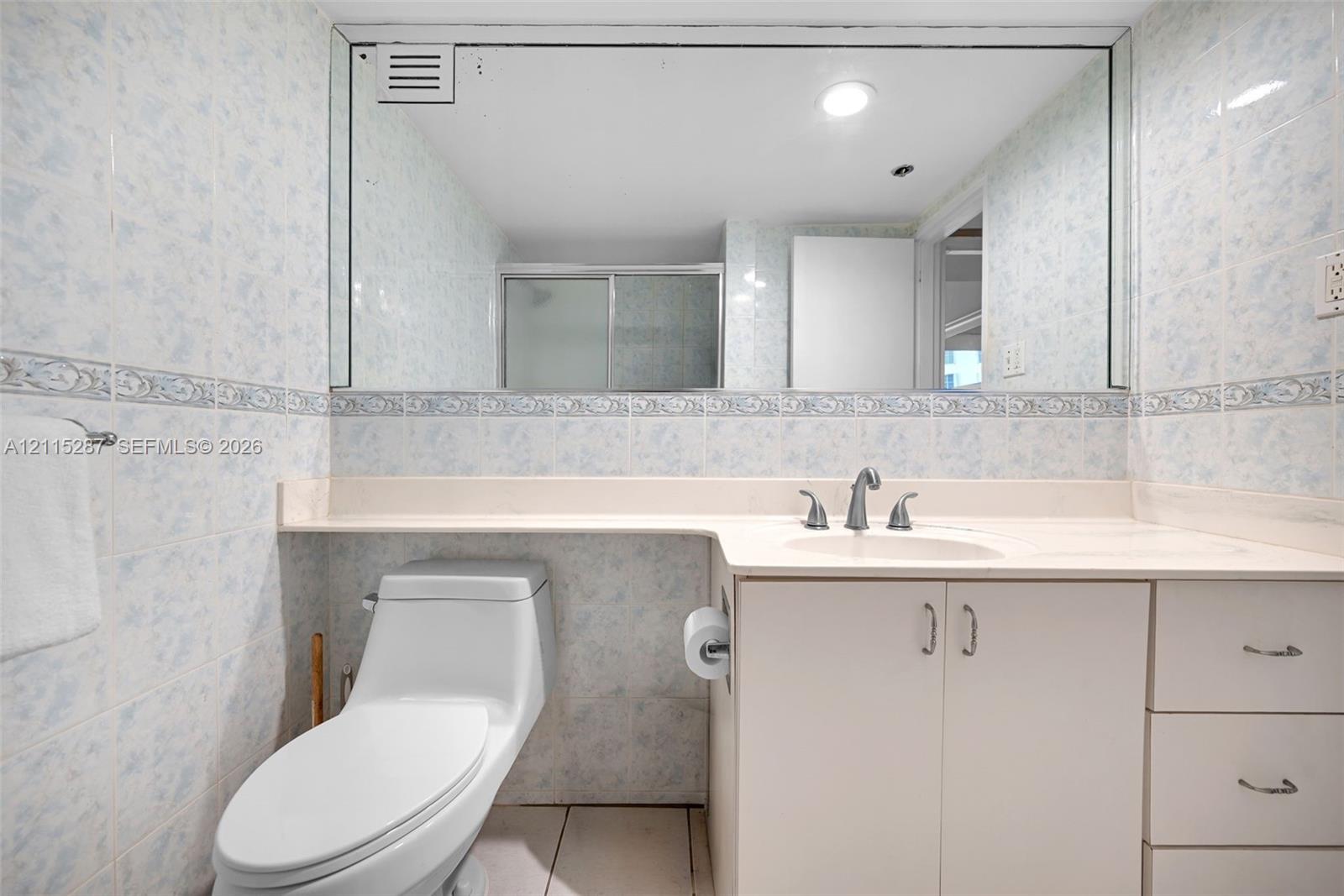 Bathroom featuring a single-basin vanity with a solid-surface countertop, floor-to-ceiling tiled walls, an expansive wall-mounted mirror, and a low-profile toilet