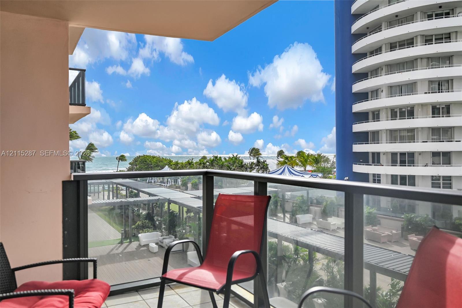 Private balcony featuring glass panel railings and tile flooring, showcasing coastal scenery and palm trees