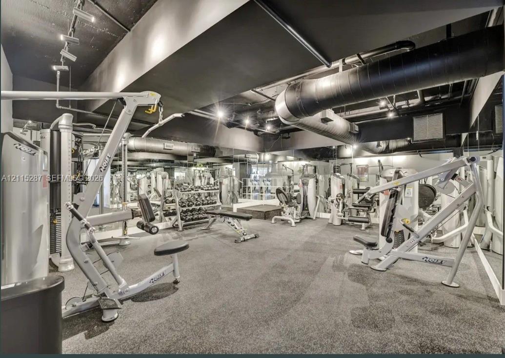 Industrial-style fitness space featuring exposed ductwork, black-painted ceiling, rubberized flooring, wall-to-wall mirrors, and track lighting