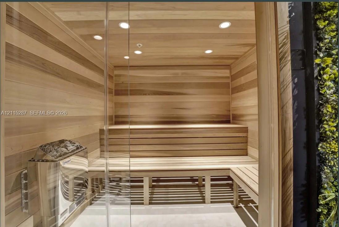 Sauna featuring horizontal wood-plank wall paneling, tiered wood-slat bench seating, recessed lighting, and a metal-encased heating unit behind a glass enclosure