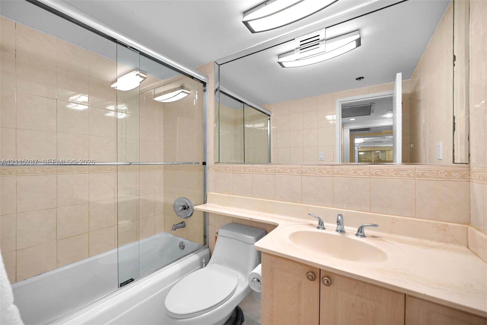 Bathroom featuring floor-to-ceiling tile surround, glass shower sliding door, vanity with integrated sink and countertop, full-width wall mirror, and dual ceiling-mounted light fixtures