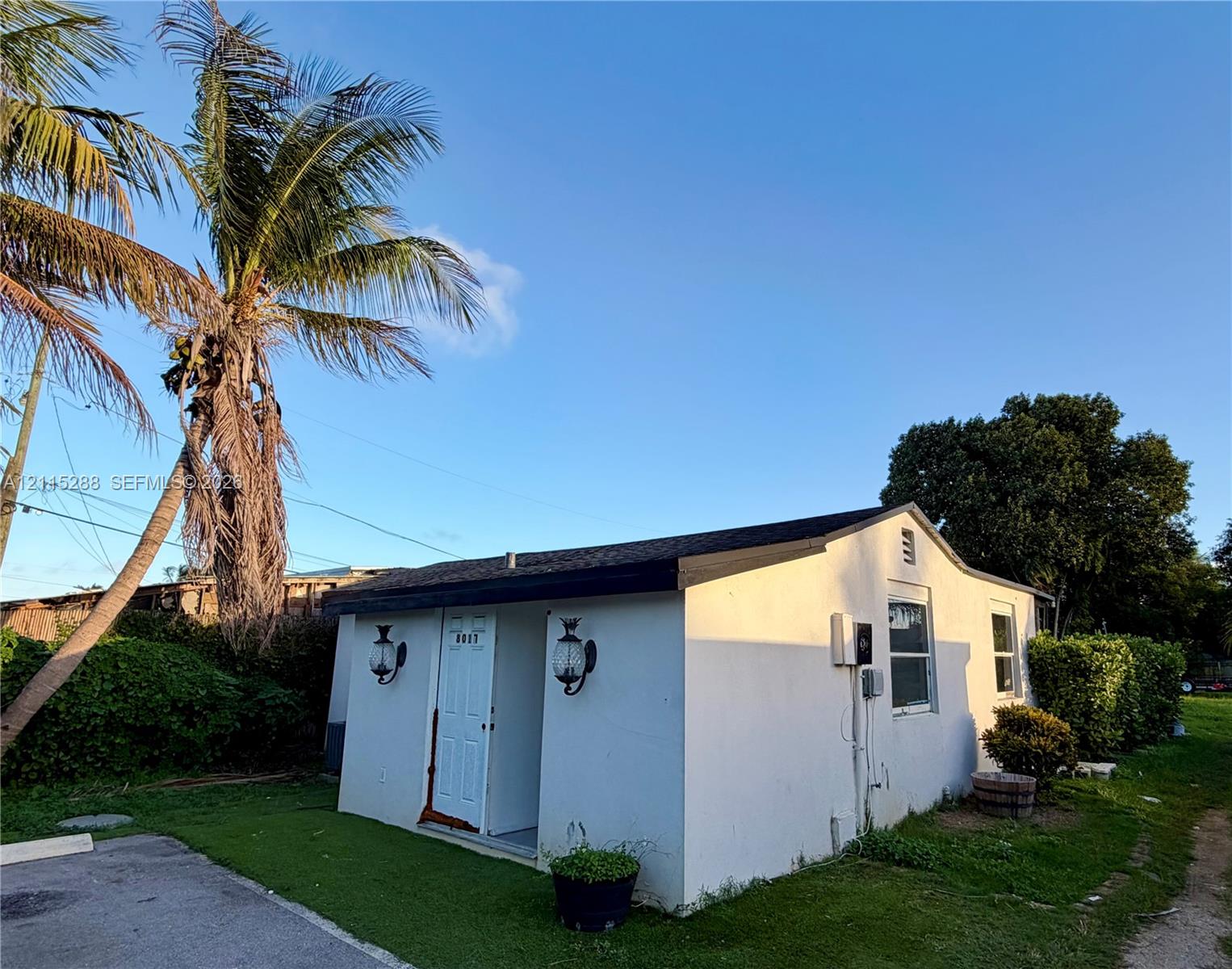 Single-story stucco exterior featuring a gabled roof, recessed entryway, wall-mounted coach lighting, and mature palm landscaping