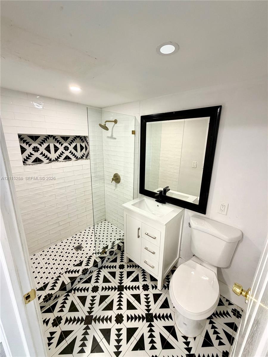 Walk-in shower with a glass panel, recessed decorative tile wall niche, geometric patterned floor tiling, white vanity cabinet, and black-framed mirror