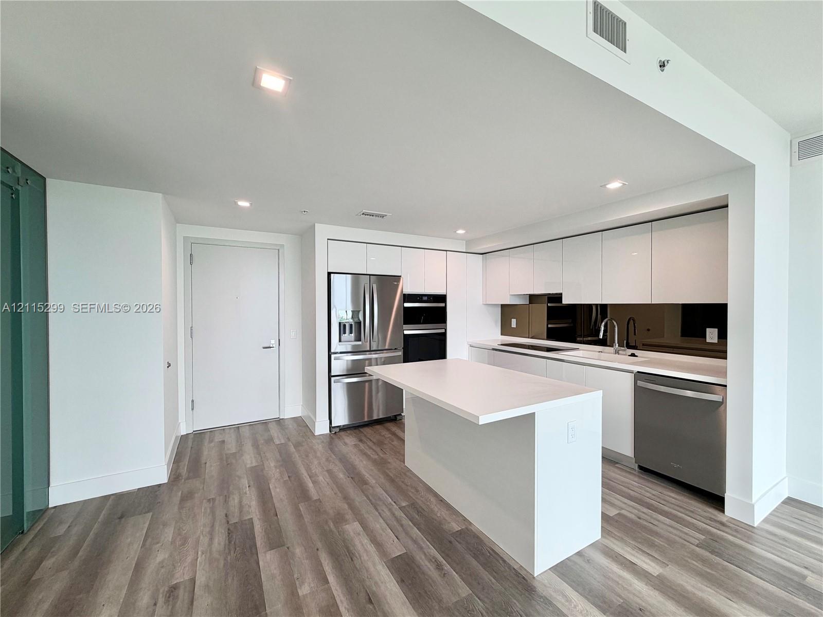 Modern kitchen featuring white flat-panel cabinetry, wood-finish flooring, stone-surfaced center island, mirrored backsplash, and stainless steel appliances