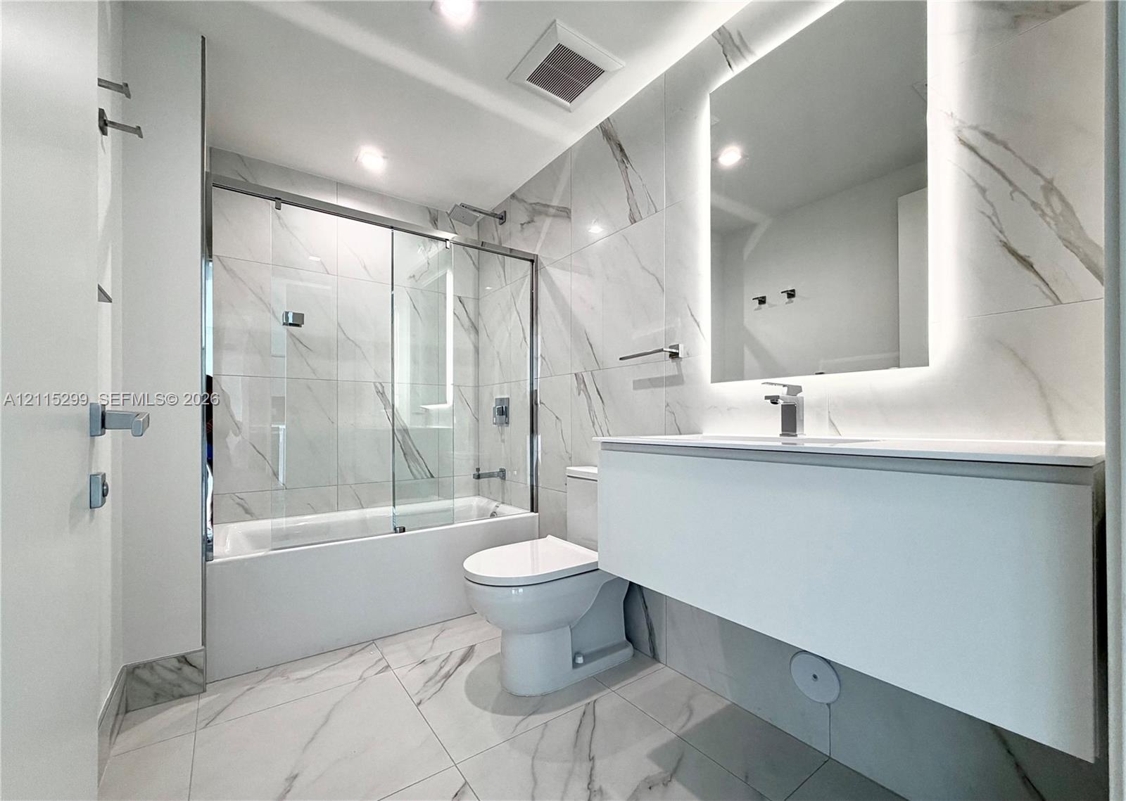 Full-height stone-patterned wall tile, floating vanity with integrated countertop, back-lit mirror, and glass-enclosed shower with polished chrome fixtures