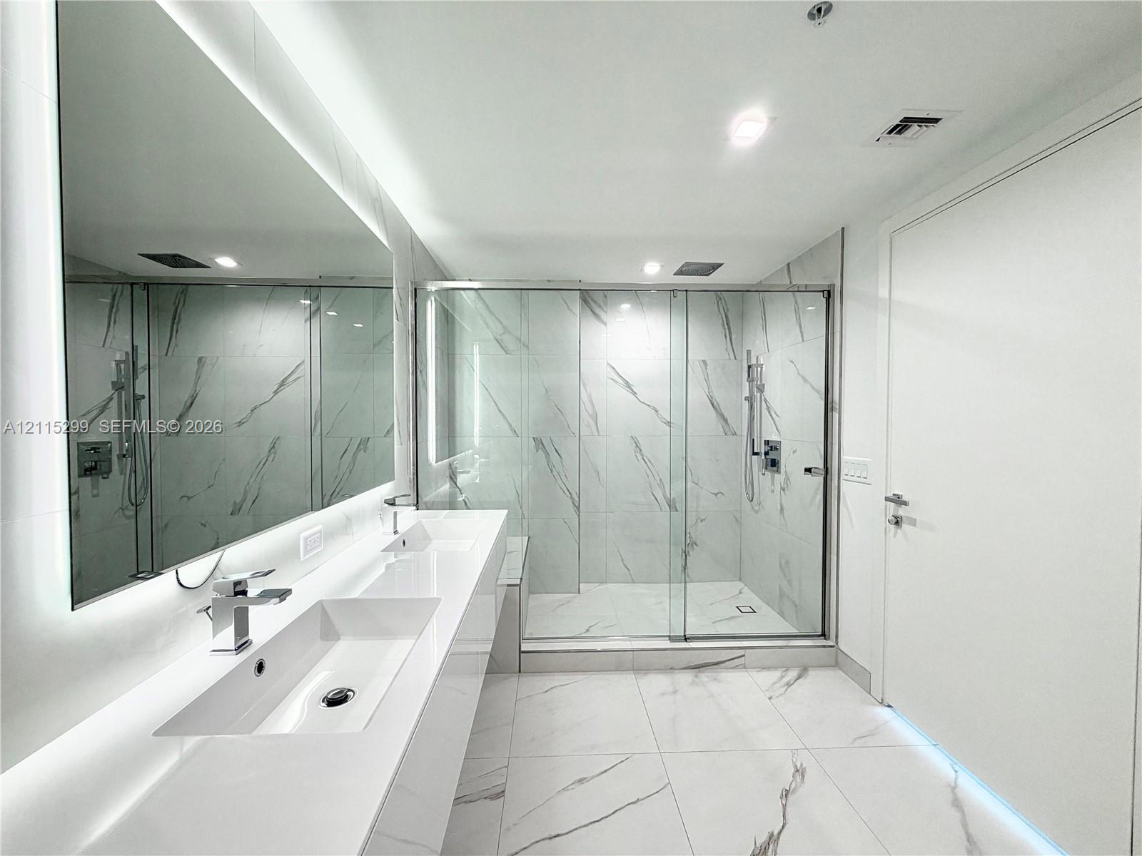 Double vanity with integrated sinks, expansive wall-mounted mirror, glass-enclosed walk-in shower with stone-finish tile surround, and large-format stone-finish floor tiling