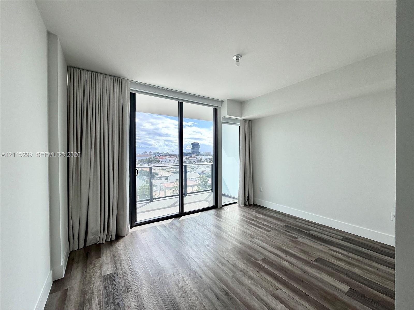 Floor-to-ceiling sliding glass door, private balcony with metal railing, wood-finish flooring, and expansive exterior urban scenery