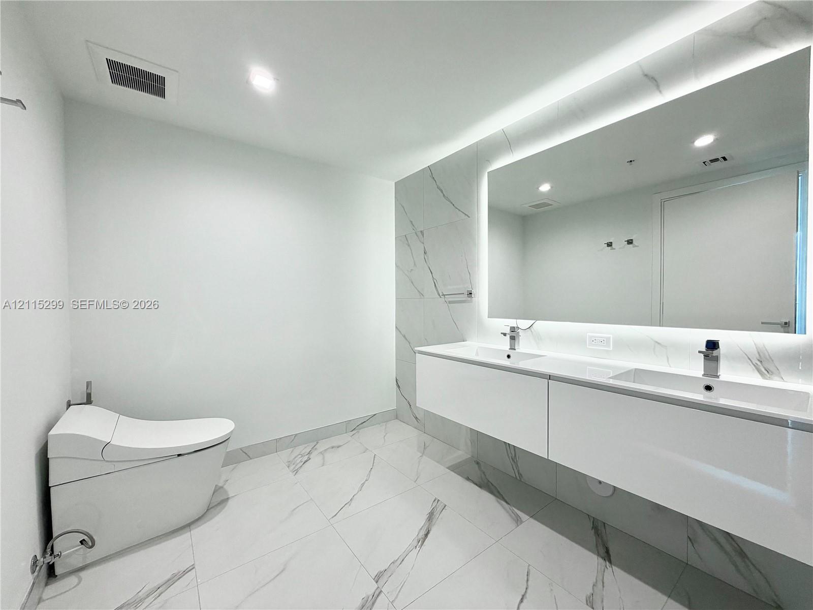 Modern floating double vanity featuring dual integrated sinks, large back-lit mirror, wall-mounted fixtures, and stone-finish floor and wall tiles