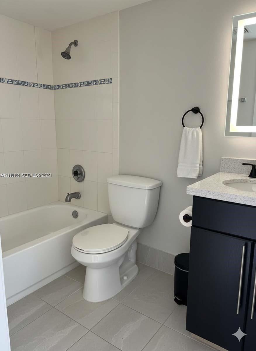Dark cabinetry with stone-finish countertop, undermount sink, matte black hardware, and an illuminated wall mirror, complemented by a tiled shower-tub combination featuring a decorative accent border