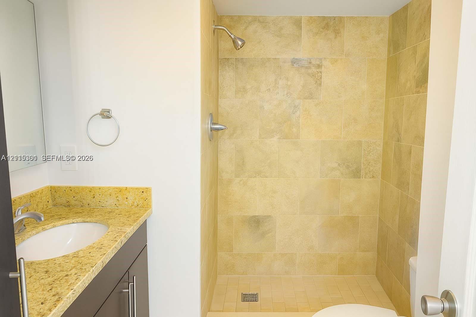Bathroom featuring a stone-finish tile walk-in shower, dark-tone vanity cabinetry, natural stone countertop, undermount sink, and chrome-finish hardware