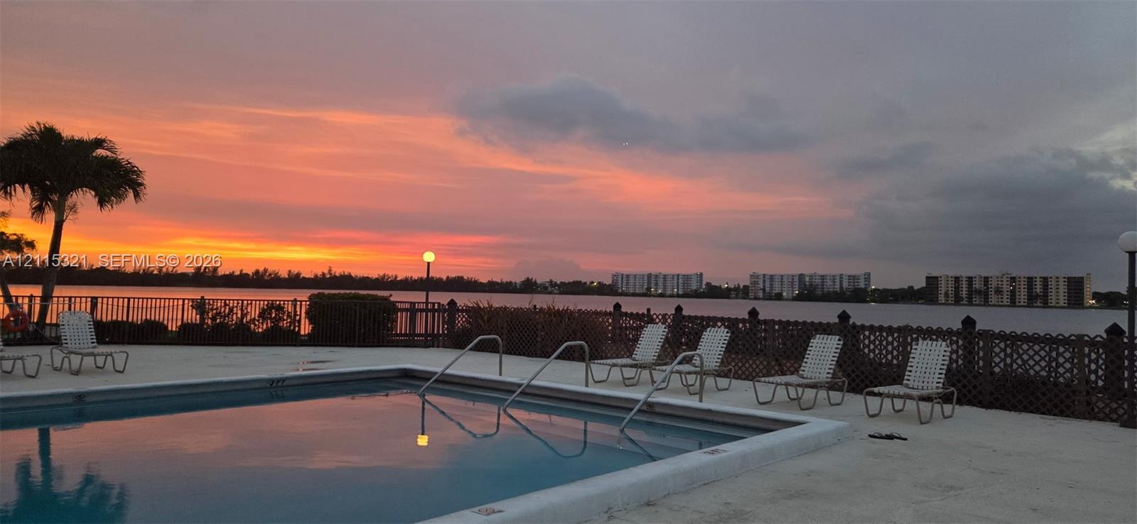 Swimming pool with a concrete deck, metal perimeter fencing, and a waterfront setting against a sunset backdrop