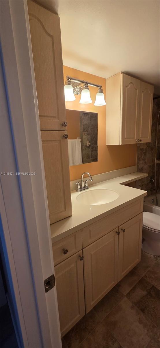 Vanity featuring light wood-finish cabinetry, a solid-surface countertop, an integrated sink, a three-light wall fixture, and overhead storage cabinetry