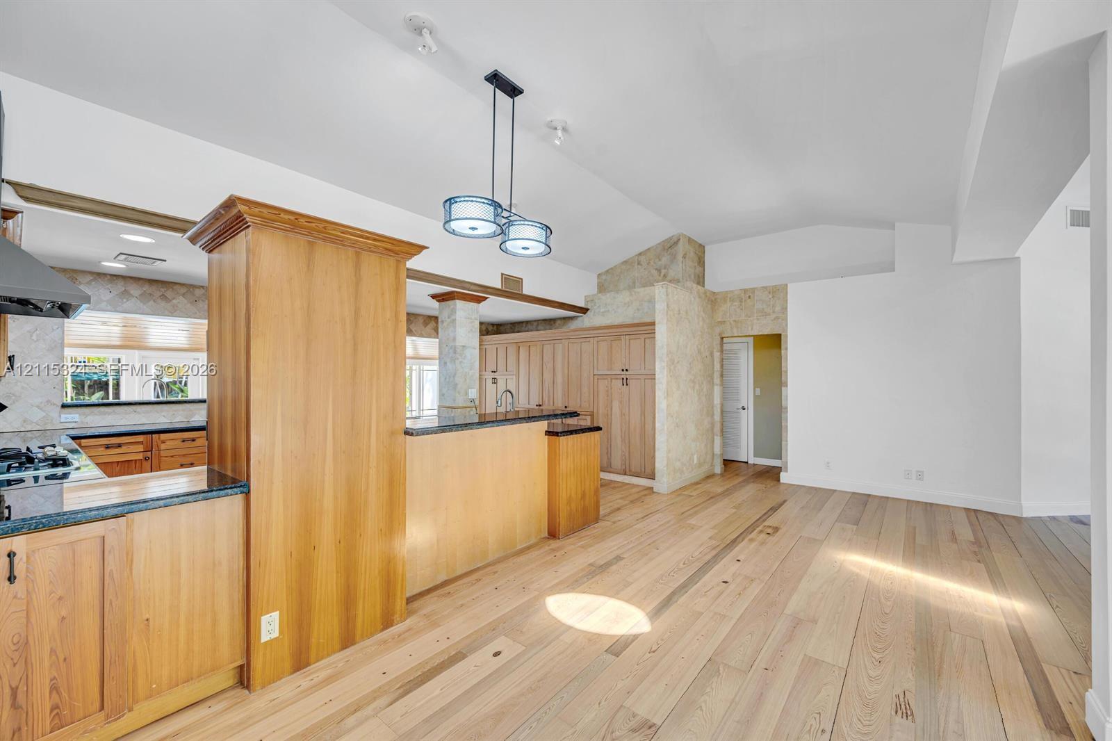 Kitchen area featuring wood-finish cabinetry, dark stone countertops, vaulted ceilings, dual-pendant lighting, and light-toned wood-finish flooring